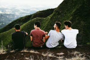 A row of four men sitting on a hiking trail in a green, lush mountain.